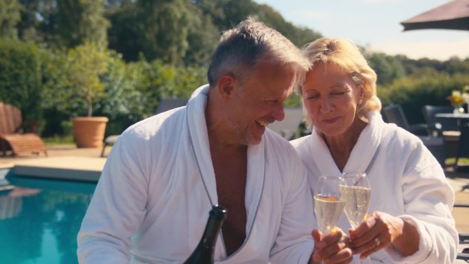 穿着长袍的老夫妇在室外的游泳池边喝香槟