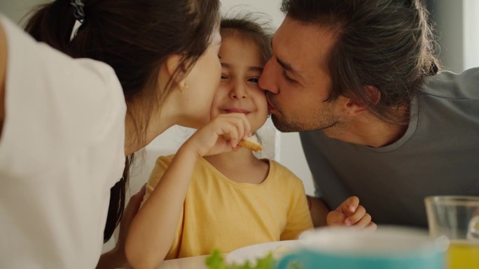 一个穿着黄色裙子的黑发小女孩正在吃早餐，她的父母在早晨的家庭餐桌上亲吻她的双颊