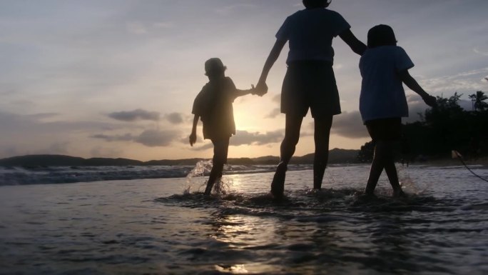 年轻的母亲带着两个儿子在日落的海滩上，夕阳的剪影母子手牵着手看着大海和日落。