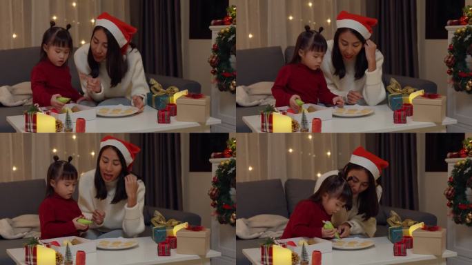 亚洲家庭圣诞快乐，新年快乐。晚上，妈妈和女儿在家里客厅的桌子上烤饼干装饰的糖霜作为圣诞礼物。
