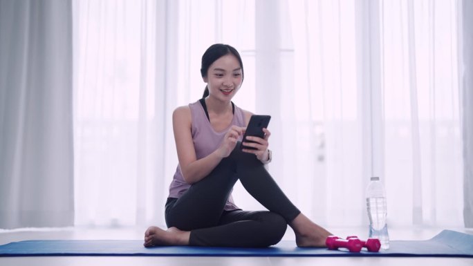 一名身着运动服的亚洲女性正在自家客厅的瑜伽垫上使用智能手机。理想的展示积极的生活方式和舒适的家庭瑜伽