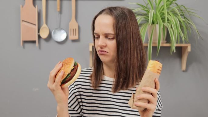 穿着条纹衬衫的棕色头发的白人女人坐在厨房里饿着肚子，手里拿着热狗和汉堡，厌恶地闻着垃圾食品。