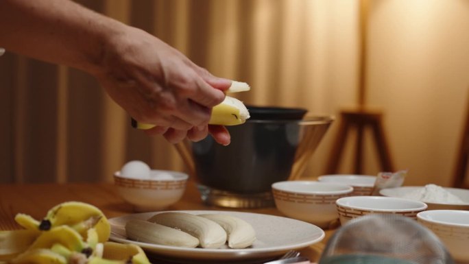 一个女人正在准备甜点，她正在剥香蕉，碗里的食材放在桌子上，温暖的灯光从一盏灯的背景。