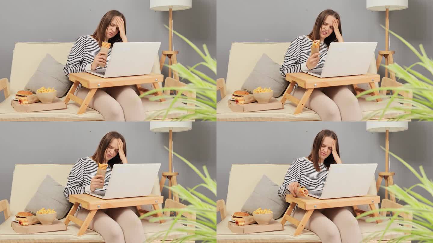 悲伤的女人带着笔记本电脑和外卖快餐坐在客厅舒适的沙发上，手里拿着热狗，无聊得头痛，没有胃口。
