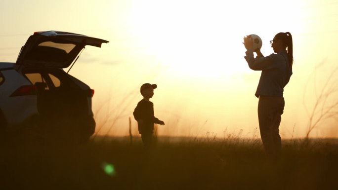 日落时分，妈妈和儿子在一辆开着后备箱的汽车旁玩球。一个幸福的家庭。夏日阳光下一家人的剪影。友好家庭的