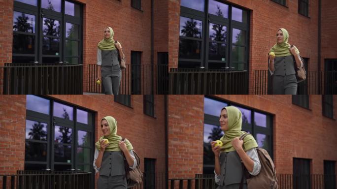 戴着头巾的美女走在城市街道上。一个穆斯林学生边走边吃苹果。伊朗、阿富汗女学生在学校大楼前。