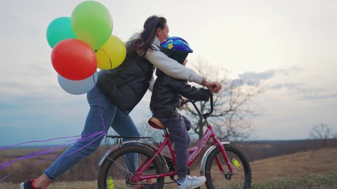 童年自行车概念。母亲教儿子骑自行车。夕阳下，年轻的妈妈第一次教儿子在乡间小路上骑自行车。快乐的孩子骑