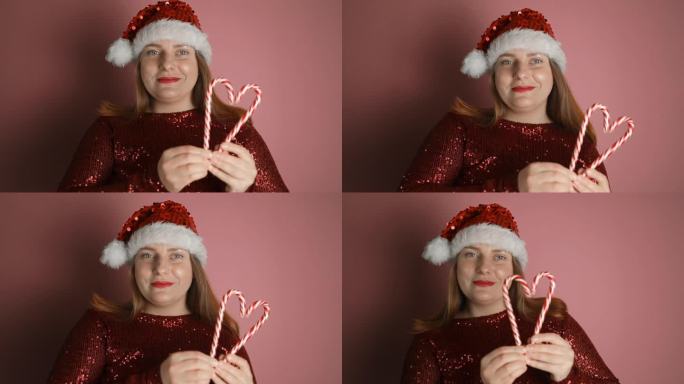 美丽可爱的女人戴着圣诞老人的帽子，穿着红色的衣服，在粉红色的背景上用糖果手杖做爱心