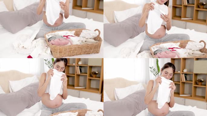 亚洲新妈妈为生第一个孩子兴奋地在家里的床上准备衣服。美丽的孕妇坐在床上，叠好宝宝的衣服，把它们分拣好