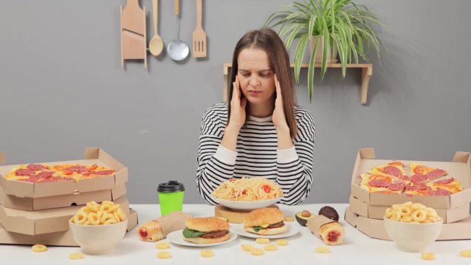 悲伤的不健康的白人妇女穿着条纹衬衫，坐在桌子上吃着各种各样的快餐，头痛，没有食欲，感到头痛。