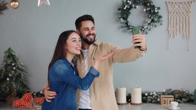 一对恩爱的夫妇用智能手机进行节日视频通话，向朋友和远方的家人致以圣诞快乐的祝福。丈夫拥抱着妻子，快乐