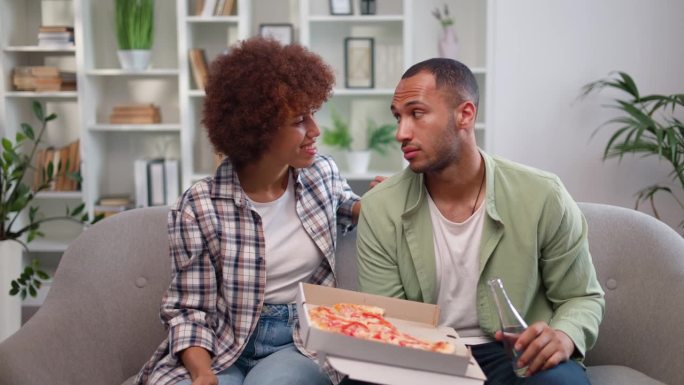 多元文化的女人手里拿着遥控器，决定看什么电视，而男朋友对她的选择完全不满意。穿着随意的夫妇正在吃披萨