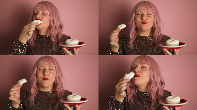 漂亮的白种人粉色头发的女人，穿着闪亮的裙子，红唇，吃着美味的奶油纸杯蛋糕，在节日里摆姿势。微笑的寿星