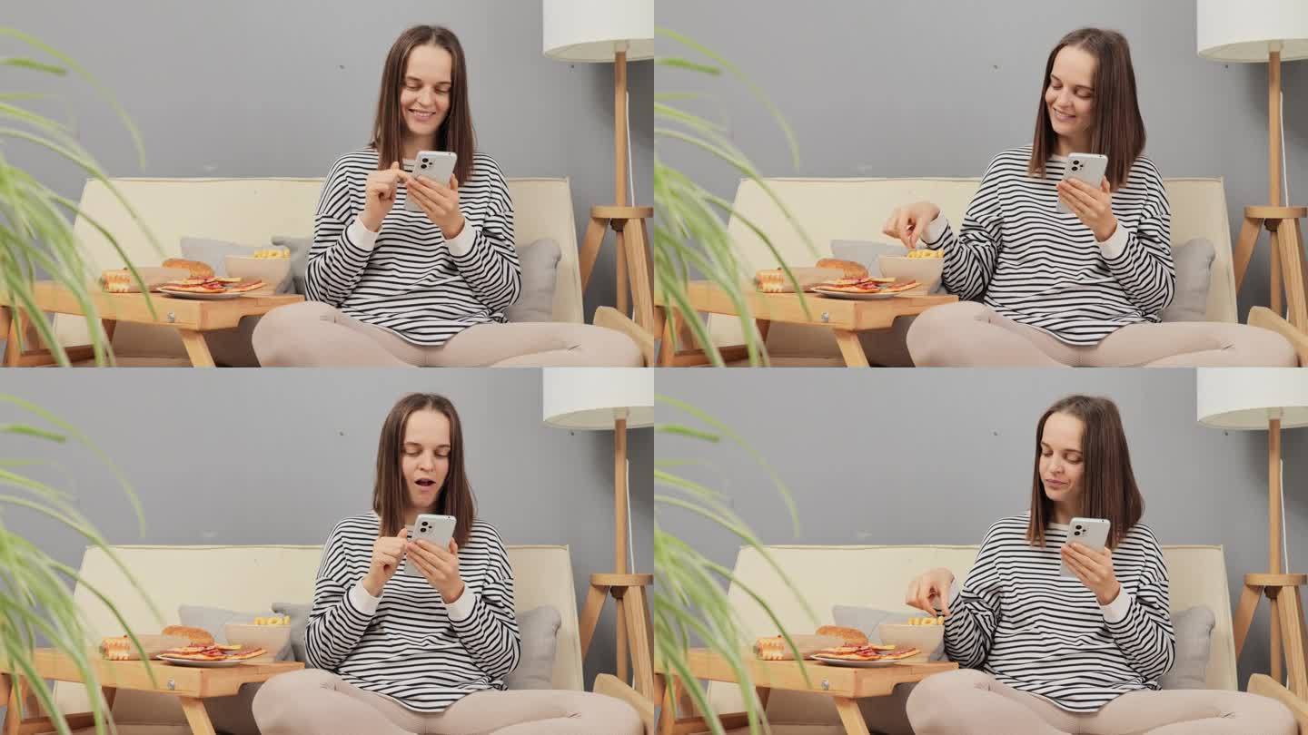 满意的女性在家放松，坐在客厅的沙发上吃洋葱零食，用手机上的应用程序在线订餐，看互联网内容吃洋葱圈。