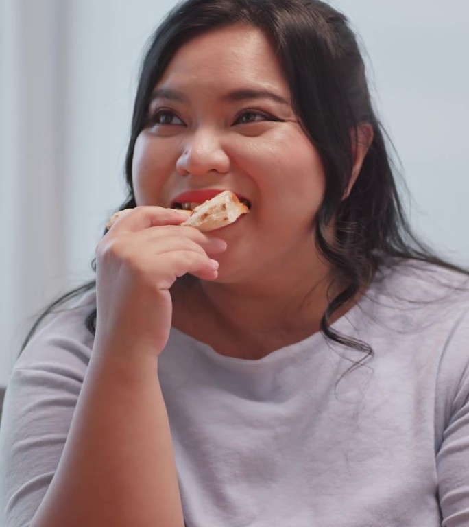垂直夹。一个胖乎乎的亚洲女人坐在家里的沙发上享受披萨。