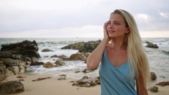 一个女人沿着沙滩漫步，蓝色的裙子在海风中飘动。金发女郎享受宁静的海滩日落，温柔的海浪拍打岩石。宁静的