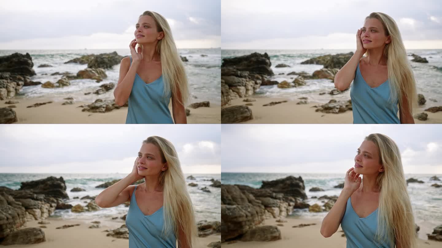 一个女人沿着沙滩漫步，蓝色的裙子在海风中飘动。金发女郎享受宁静的海滩日落，温柔的海浪拍打岩石。宁静的