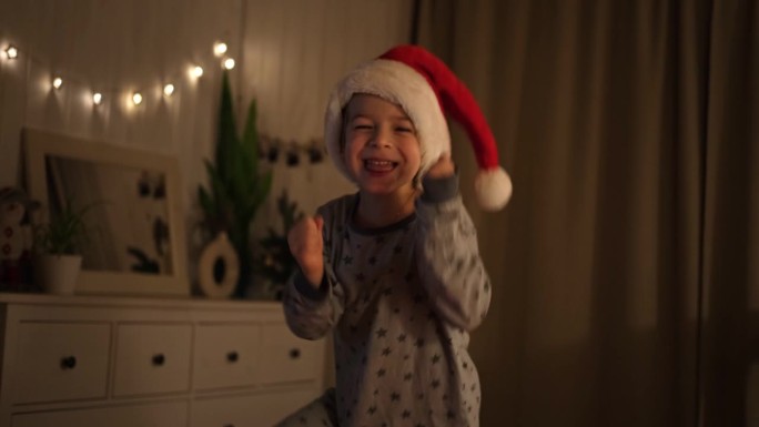 圣诞快乐。滑稽的小男孩边笑边拿着圣诞礼盒，玩得很开心。新年快乐。兴奋的小孩穿着睡衣，在床上跳来跳去。