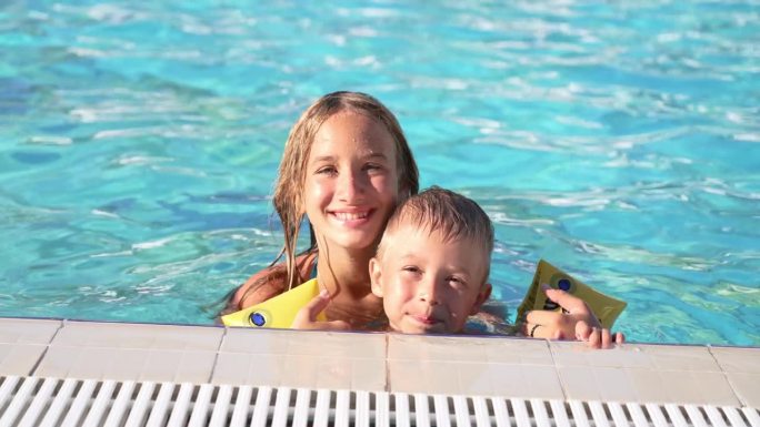 快乐的孩子们在游泳池游泳的特写。哥哥和妹妹。年轻漂亮的女孩和小男孩开怀大笑。戴着充气臂章的男孩抓住泳