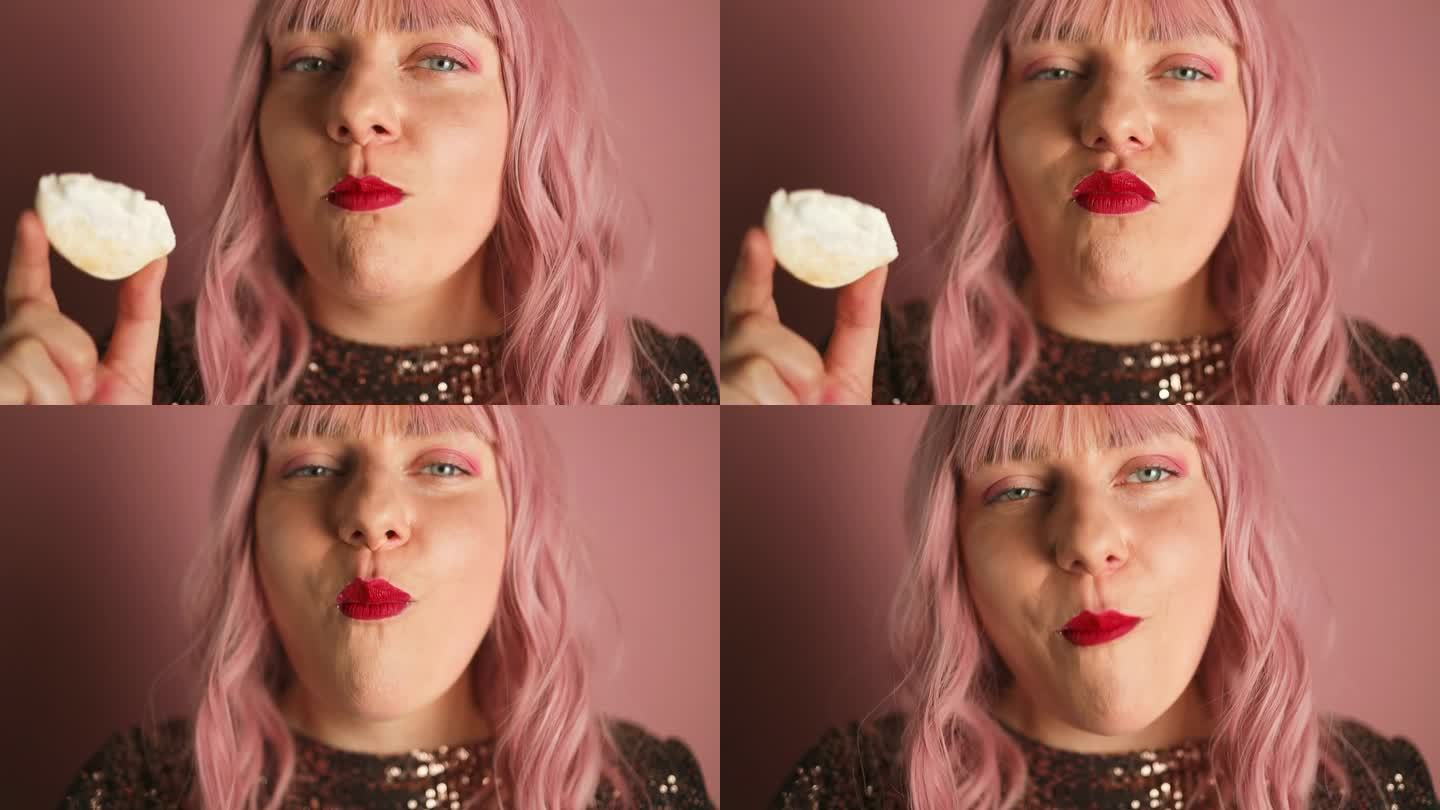 快乐的白种人粉色头发的女人，穿着闪亮的裙子，红唇，吃着美味的奶油纸杯蛋糕，在节日里摆姿势。微笑的寿星