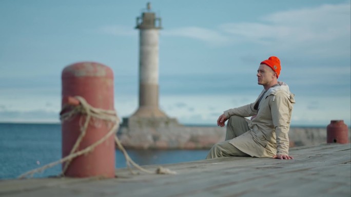 渔民独自坐在老灯塔附近的码头上，年轻的成年男子穿着运动服，戴着红帽子