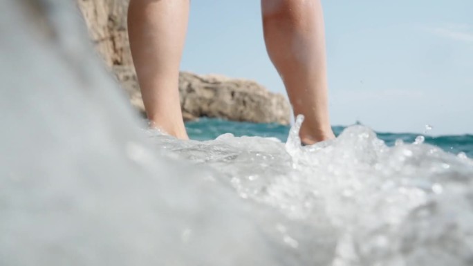 低角度拍摄的女性站在岸边的岩石上，双脚被海浪覆盖的慢镜头。