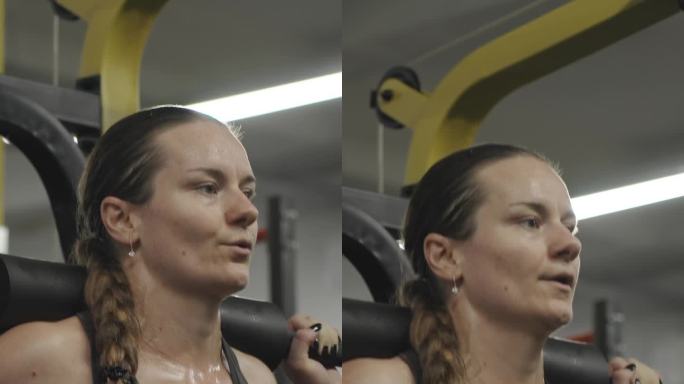 垂直视频。一个留着长辫子的年轻女子在史密斯健身机上做深蹲的特写。她满头大汗，把杠铃放回原位。