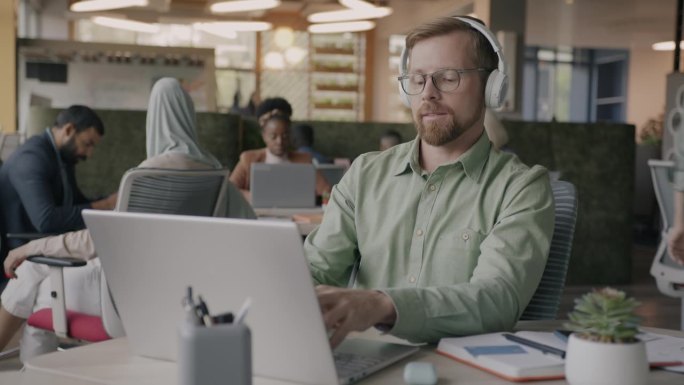 快乐的年轻员工在联合办公中心戴着耳机听音乐，使用笔记本电脑