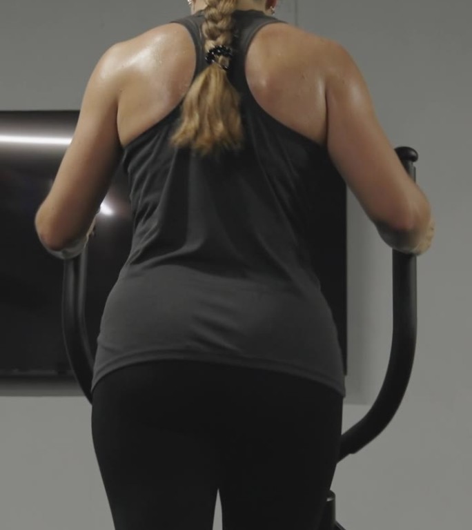 垂直视频。一个长头发扎成辫子的女人在椭圆机上锻炼，她的背汗流浃背。