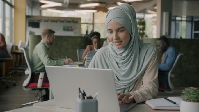 戴着头巾的穆斯林妇女用笔记本电脑在开放式办公室里享受创造性的活动