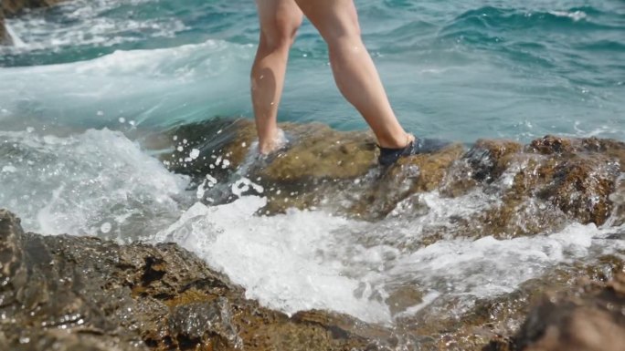 一个穿着泳装的年轻女子站在海边的岩石峭壁上，海浪冲刷着她的脚。缓慢的运动。