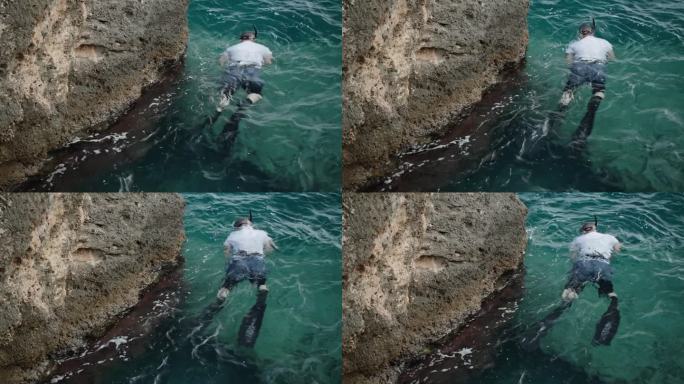 一个戴着鱼鳍和面具的男人沿着海边的悬崖游泳，捕鱼。他腰上系着一条捕获的鱼。