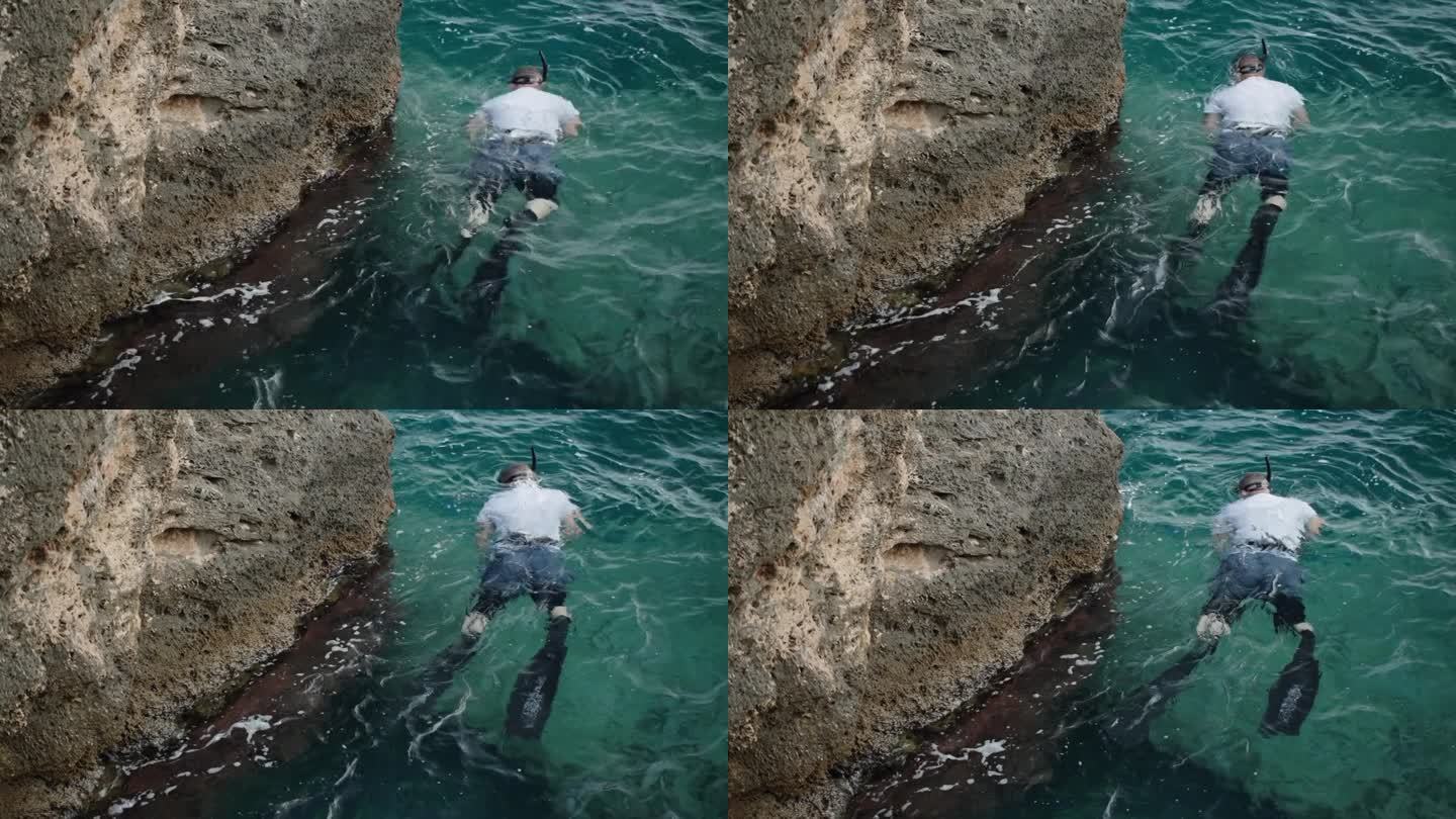 一个戴着鱼鳍和面具的男人沿着海边的悬崖游泳，捕鱼。他腰上系着一条捕获的鱼。