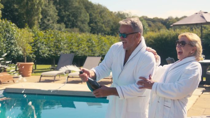 度假的老夫妇穿着长袍，在酒店游泳池边开瓶喷香槟