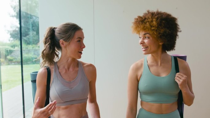 两个女性朋友穿着运动服，带着运动垫去健身房或瑜伽室锻炼