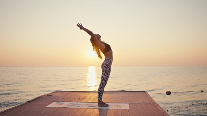 一个年轻的金发女孩穿着黑白相间的裤子和黑色上衣，在一个特殊的运动垫子上做瑜伽，做半月形练习，在日出时