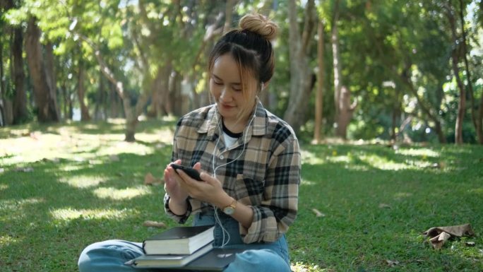 一位年轻女子坐在公园里用手机，戴着耳机听音乐。
