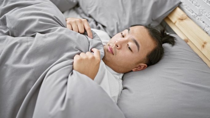 英俊，年轻的亚洲男子，惊恐的表情明显，躺在床上盖着毯子，害怕的早晨醒来