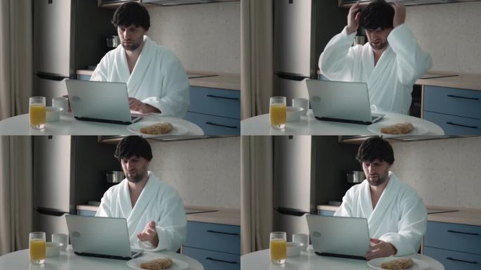 一个没刮胡子、黑头发、穿着浴袍的男人正在厨房里用笔记本电脑工作，对未能达成的协议感到失望