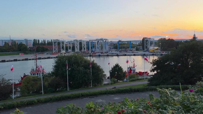 码头上的日落景象，停泊着船只和工业建筑，旗帜在风中飘扬，还有柔和的天空——Westerplatte 