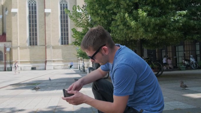 一名游客用手机蹲在城市广场上，通过手机上的在线应用程序预订酒店。男性旅客想在最后一刻订票