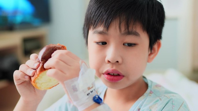 一个年轻的亚洲男孩坐在医院的病床上，品尝着一种简单但营养丰富的面包小吃。尽管他正在住院治疗，但这个孩