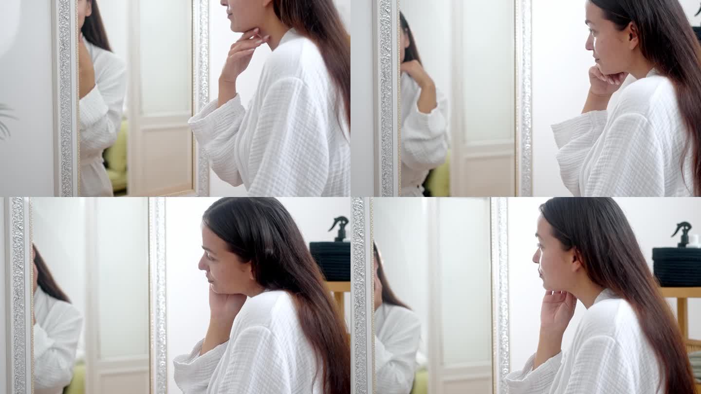一个女人，穿着浴袍，在家里练习美容护肤程序，对着浴室的镜子审视自己