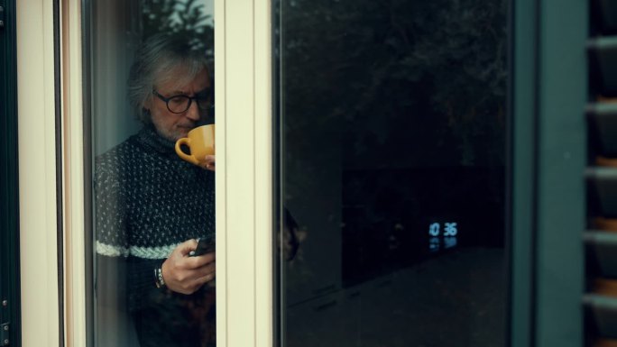 一名成年男子一边喝着热饮，一边在窗边看手机