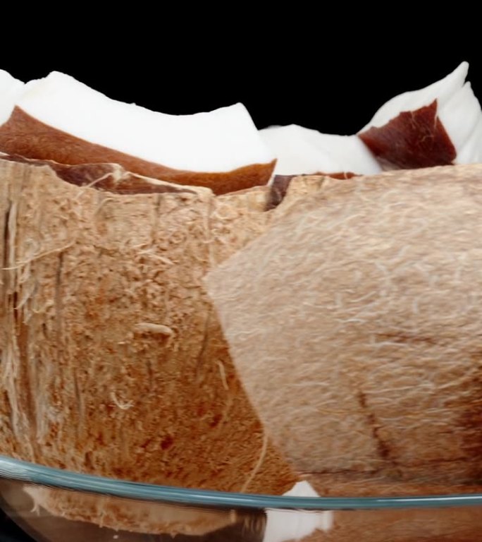 垂直视频。一个棕色的椰子碎成碎片，白色的果肉放在一个玻璃盘子里。它在一个孤立的背景上循环旋转。