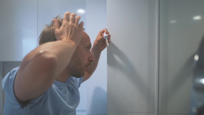 男人用抗脱发精华液或产品，从移液管上滴到浴室里的秃顶上，看着镜头