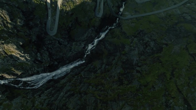 无人机飞过绿色的岩石峡湾表面和狂野的河流，显示史诗山蜿蜒的道路，由阳光覆盖