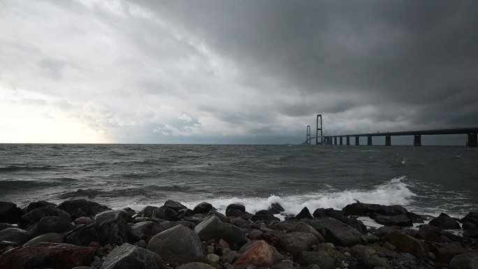 丹麦连接两个海岸的大腰带大桥的航拍画面。在横跨地平线的Storebælt桥前，巨浪冲击着石头。
