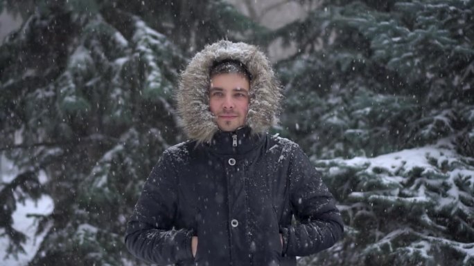 冬天下大雪，一个年轻人站在冷杉树的背景下。一名身穿羽绒服、戴着兜帽的男子站在那里看着镜头。缓慢的运动