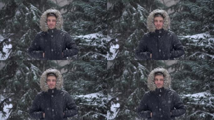 冬天下大雪，一个年轻人站在冷杉树的背景下。一名身穿羽绒服、戴着兜帽的男子站在那里看着镜头。缓慢的运动